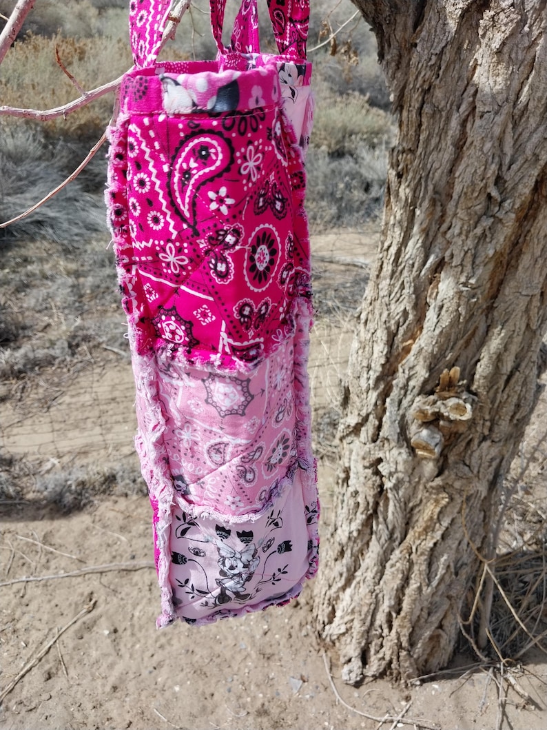 Puede incluir: Un bolso tote rosa hecho a mano con un dise&ntilde;o de patchwork. El bolso presenta varios patrones de cachemira y florales en tonos de rosa, blanco y negro. El bolso est&aacute; colgado de una rama de &aacute;rbol.