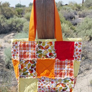 May include: A quilted tote bag featuring a patchwork design of autumn-themed fabrics in shades of orange, yellow, red, and white. The bag has two bright orange fabric handles and a square-patterned construction with frayed seams between squares.