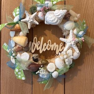 May include: A round wreath decorated with seashells, starfish, and ribbons in shades of white, beige, green, and blue. The word "welcome" is displayed in the center. The wreath is hanging on a wooden door.