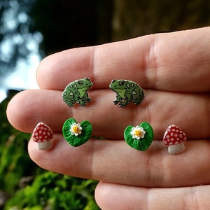 May include: A set of six earrings featuring nature-inspired designs. Two green frog earrings, two green lily pad earrings with white flowers, and two red and white mushroom earrings.