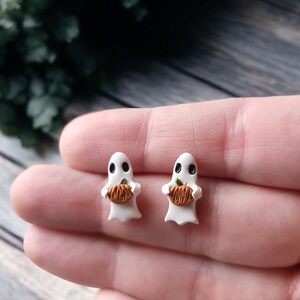 May include: A pair of white ghost earrings with black eyes and a small orange pumpkin in their hands.