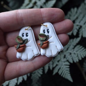 May include: A pair of white ghost-shaped earrings, each featuring a small green plant in a brown pot held by the ghost. The earrings have gold-toned metal jump rings at the top for attachment, with smooth, matte surfaces and defined facial features.