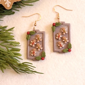 May include: Gingerbread man earrings with gold-colored hooks. Each earring features a rectangular design with several miniature gingerbread men, holly leaves, and red berries. Festive holiday jewelry.