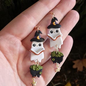 May include: A pair of Halloween-themed earrings. Each earring features a white ghost wearing a black witch hat with a crescent moon, holding a book, and a black cauldron with green bubbles and a gold star charm.