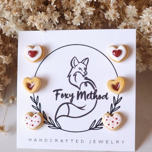 May include: Set of four heart-shaped cookie earrings. The cookies are decorated with red and white icing and sprinkles. The earrings are displayed on a white card with the text "Foxy Method Handcrafted Jewelry".