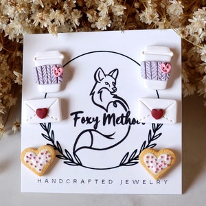 May include: A set of three handmade polymer clay earrings featuring a coffee cup, a letter, and a heart-shaped cookie. Each earring has a pink heart detail. The earrings are displayed on a white card with the text "Foxy Method" and "Handcrafted Jewelry".