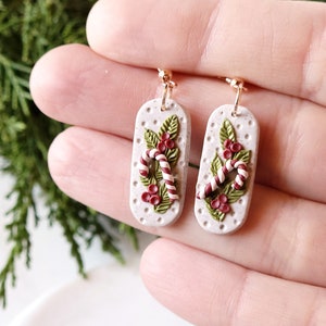 May include: A pair of white dangle earrings with a Christmas theme. The earrings feature a candy cane, holly leaves, and red berries.