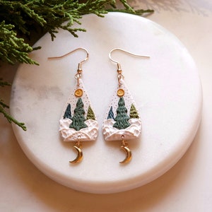 May include: A pair of white triangle-shaped earrings with a gold crescent moon charm. The earrings feature a winter scene with green trees and a yellow sun.