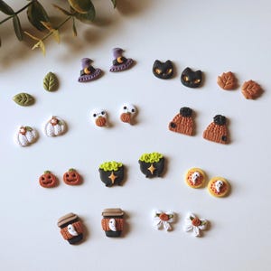 May include: A collection of Halloween-themed miniature charms. The charms include witch hats, black cats, pumpkins, ghosts, cauldrons, coffee cups, leaves, and bows. The charms are in various colors, including orange, black, white, and purple.