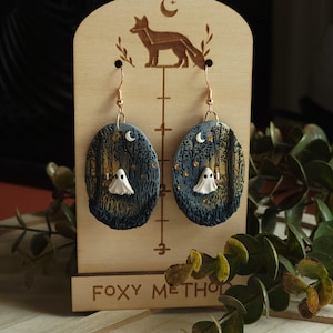 May include: Handmade oval earrings depicting a forest scene with a white ghost and a crescent moon. The earrings are displayed on a wooden stand with a fox silhouette and the text "FOXY METHOD". The earrings have gold-coloured hooks.