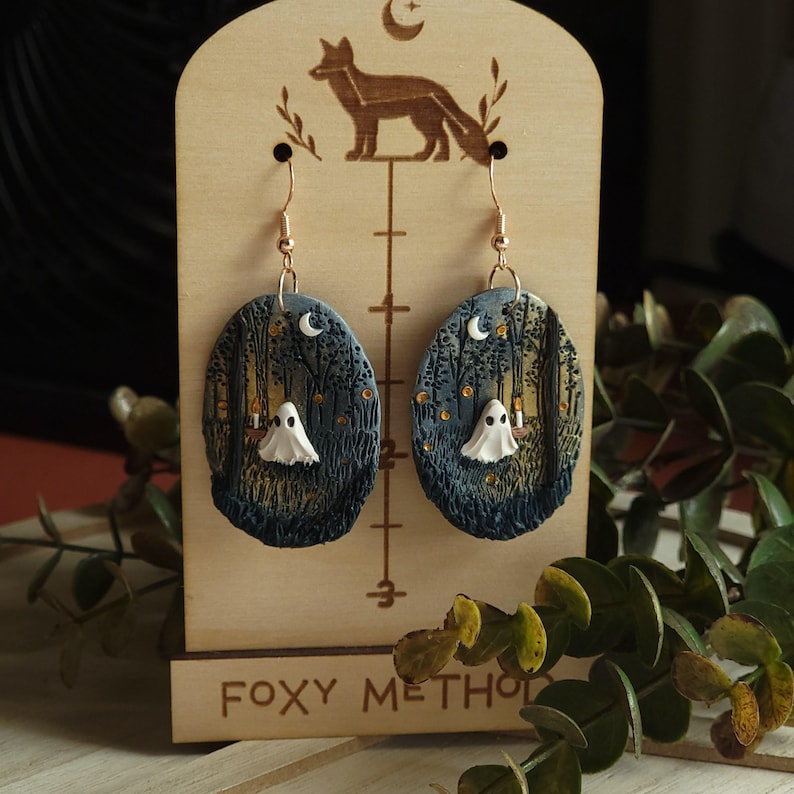 May include: Handmade oval earrings depicting a forest scene with a white ghost and a crescent moon. The earrings are displayed on a wooden stand with a fox silhouette and the text "FOXY METHOD". The earrings have gold-coloured hooks.