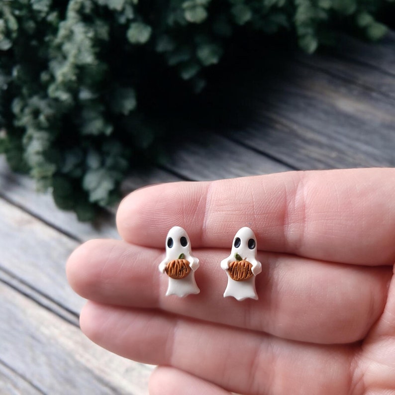 May include: A pair of white ghost earrings with black eyes and a small orange pumpkin in their hands.