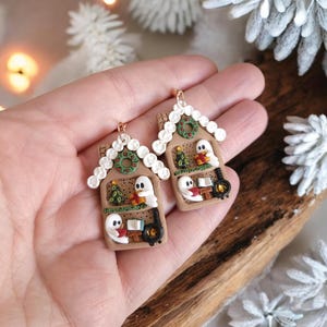 May include: A pair of festive gingerbread house earrings. Each earring features a brown house with white icing details, a green wreath, and a ghost figure. The houses have a Christmas tree, a fireplace, and a ghost holding a gift.