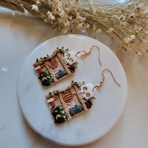 K&ouml;nnte beinhalten: Ein Paar goldene Ohrh&auml;nger mit Halloween-Motiv. Jeder Ohrring zeigt ein Miniatur-Lehmhaus mit einem Sch&auml;del, Ranken und anderen gruseligen Details.