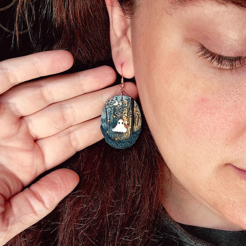 May include: Oval earring with a copper hook, depicting a Halloween forest scene. The design includes a white ghost, trees, and a moon, set against a backdrop of blues, greens, and yellows. The earring has a whimsical and spooky aesthetic.