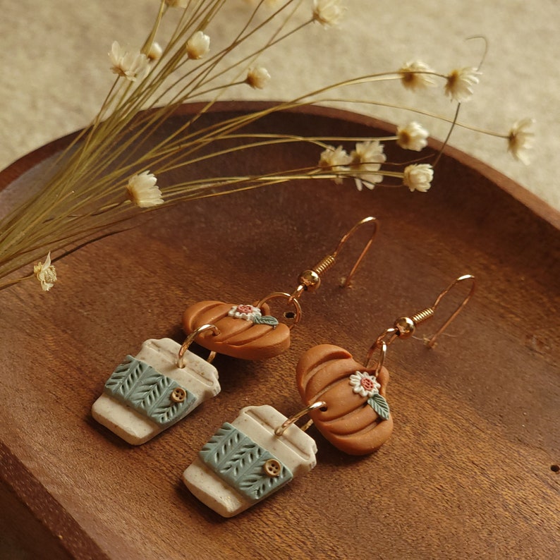 Op de afbeelding: Een paar gouden oorhangers met een witte en groene koffiebeker charme en een bruine pompoen charme met een witte bloem. De charmes zijn gemaakt van polymer clay.
