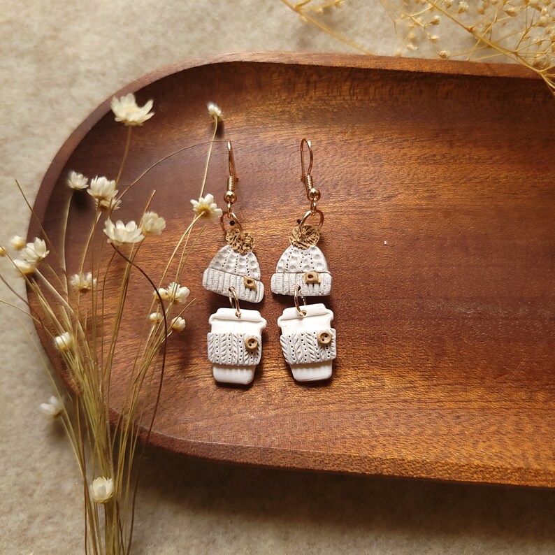 May include: A pair of white ceramic earrings with a coffee cup and a knit hat design. The earrings are on a wooden surface.