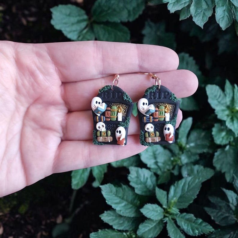 May include: A pair of black and gold earrings shaped like tombstones with ghosts, skulls, and books. The tombstones have the word "RIP" on them.