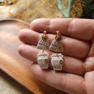 May include: A pair of dangle earrings featuring a white ceramic coffee cup with a knitted pattern and a white ceramic beanie with a gold pom-pom.
