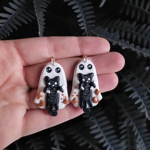 May include: A pair of Halloween-themed earrings. Each earring features a white ghost with black eyes and a black cat. The cat is holding its arms out. There are small orange stars and crescent moons on the ghost's cloak. The earrings have gold-colored hooks.