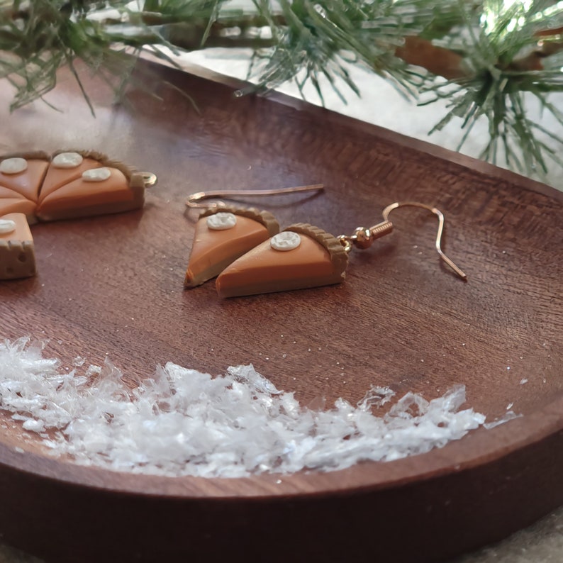 May include: A pair of gold dangle earrings with a miniature pumpkin pie slice charm. The earrings are displayed on a wooden tray with white glitter.