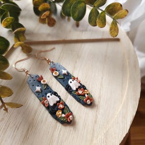 May include: A pair of dangle earrings with a dark blue background, white ghosts, orange and green flowers, and gold accents.