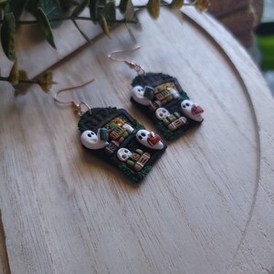 May include: A pair of black and green earrings shaped like a bookshelf with ghosts and books. The earrings are made of polymer clay and have gold hooks.