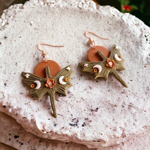 May include: A pair of green dragonfly earrings with a brown sun and moon accents. The earrings are made of polymer clay and have a whimsical, bohemian style.