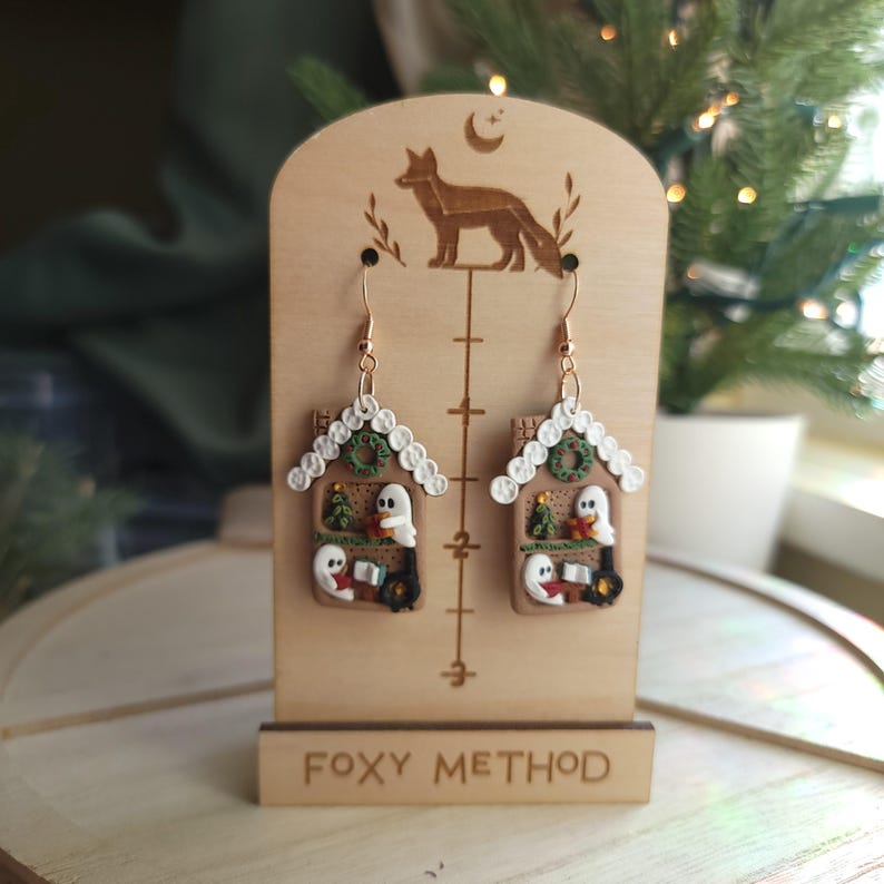 May include: A pair of whimsical house-shaped earrings with a Christmas theme. The earrings feature a brown house design with white trim, a green wreath, and a ghost figure. The earrings are displayed on a wooden stand with the text "Foxy Method".