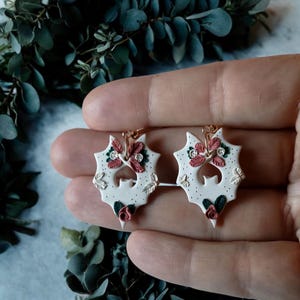 May include: A pair of white ceramic earrings shaped like bats with a floral design. The earrings have pink and green flowers and gold accents.