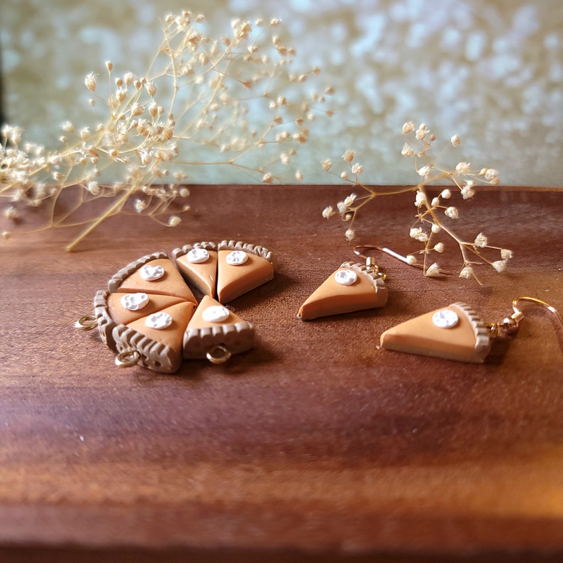 May include: A set of handmade polymer clay earrings featuring miniature pumpkin pie slices with white frosting and a pearl-like detail. The earrings are displayed on a wooden surface with dried flowers.