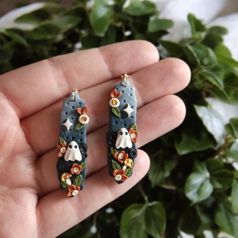 May include: A pair of dangle earrings with a dark blue background and white ghost figures. The earrings are decorated with orange, yellow, and black flowers and mushrooms.