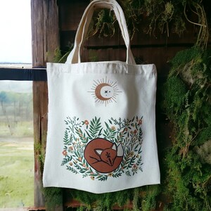 May include: A white canvas tote bag with a design of a sleeping fox surrounded by flowers and greenery. The design also includes a sun and moon with a star in the center.