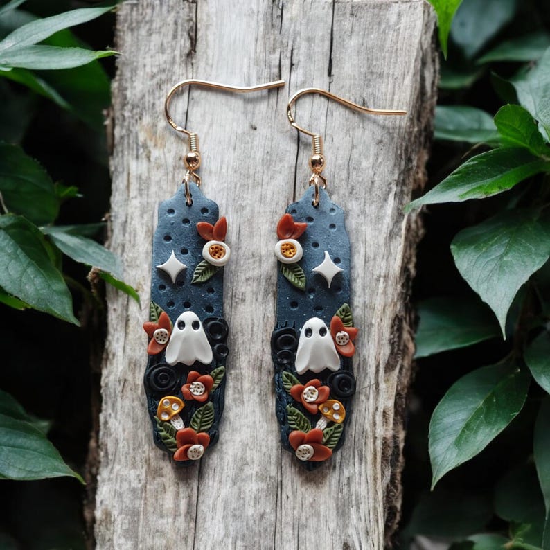 May include: A pair of dangle earrings with a dark blue background, white ghosts, orange and white flowers, and gold accents.