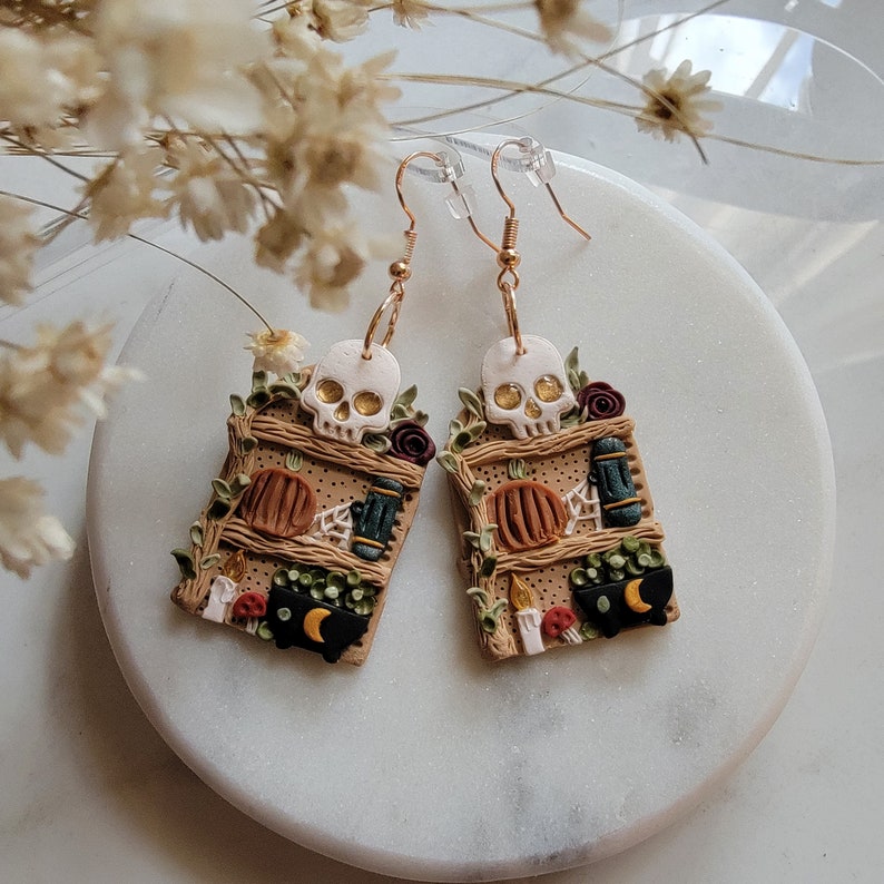 K&ouml;nnte beinhalten: Ein Paar goldene Ohrringe mit Halloween-Motiv. Jeder Ohrring zeigt ein Miniatur-Holzregal mit einem Sch&auml;del, K&uuml;rbissen, Kerzen und anderen gruseligen Details.
