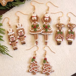 May include: A collection of festive Christmas earrings. The earrings feature gingerbread men, nutcrackers, ballet dancers, reindeer, and Christmas trees. Each earring is brown, white, and red with gold-colored hooks. The earrings are arranged on a white surface.