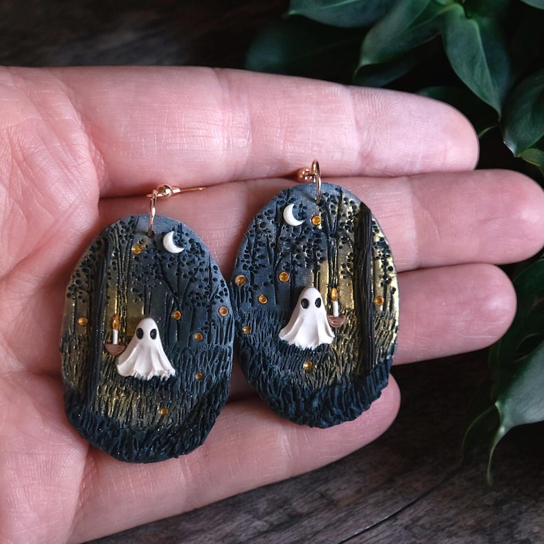 May include: Oval earrings depicting a spooky forest scene with a white ghost holding a candle. The earrings have a gold background with a crescent moon and gold accents. The earrings are held in a hand.