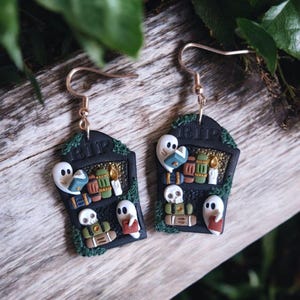 May include: A pair of black and green Halloween-themed earrings featuring a tombstone with the inscription "RIP" and ghosts reading books. The tombstone is decorated with skulls, candles, and books.