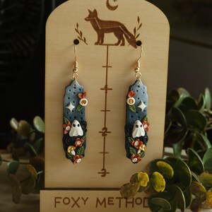 May include: A pair of dangle earrings with a blue and white ghost design. The earrings feature a ghost with a white face and black eyes, surrounded by orange and white flowers. The earrings are hanging from gold hooks.