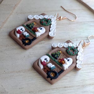 May include: A pair of handmade gingerbread house earrings. Each earring features a brown rectangular base with white frosting accents, a green wreath, and ghost figures. The earrings have gold-colored ear wires.