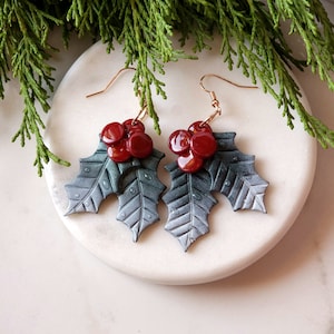 May include: A pair of silver and green holly leaf earrings with red berry accents. The earrings are made of polymer clay and have a delicate, festive design.