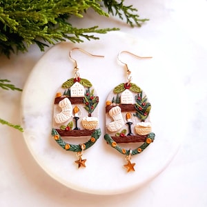 May include: A pair of gold dangle earrings with a white, brown, and green clay charm depicting a winter scene with a house, a stocking, a candle, and a bowl. The charm is attached to a gold star.