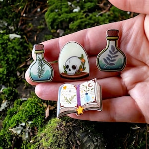 May include: A set of four witchy themed enamel pins featuring a skull in a glass dome, a book with a crescent moon and a potion bottle, and two potion bottles with leafy sprigs.