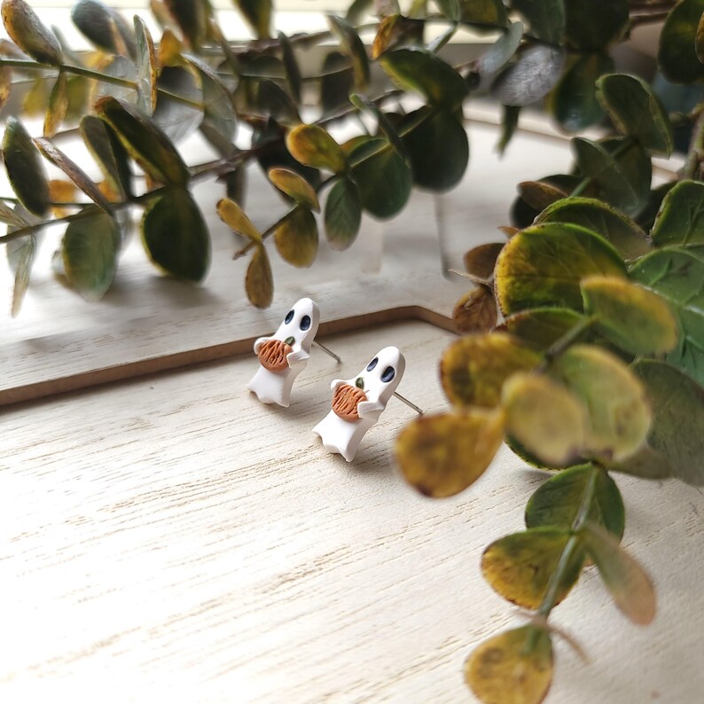 May include: A pair of white ghost earrings with black eyes and a small orange pumpkin in their hands. The earrings are on a white wooden surface.