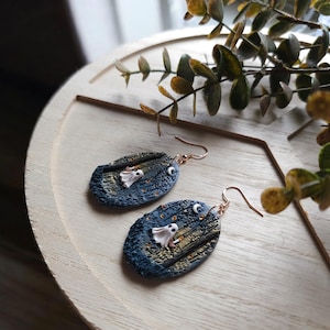 May include: Handmade oval earrings depicting a spooky night scene. Each earring has a dark blue background with a white ghost, a crescent moon, and gold accents. The earrings have copper-coloured ear wires.