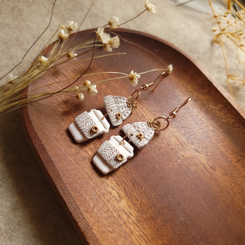 May include: A pair of white ceramic earrings with a coffee cup and a winter hat design. The earrings are on a wooden surface.