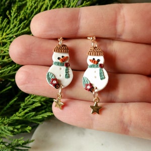May include: A pair of dangle earrings featuring a white snowman with a brown hat, green scarf, and red flower accents. Each snowman is attached to a gold star charm.