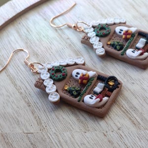 May include: A pair of festive gingerbread house earrings. The earrings are brown with white icing accents, a green wreath, and small gift boxes. The earrings have gold-colored ear wires.