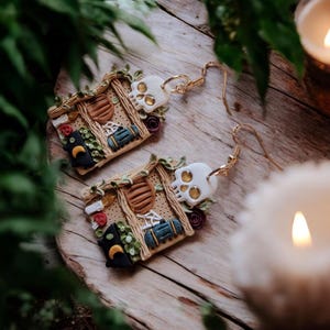 K&ouml;nnte beinhalten: Ein Paar handgefertigte Ohrringe aus Polymer Clay, die ein Miniaturhaus mit einem Sch&auml;del, einer schwarzen Katze und anderen Details zeigen. Die Ohrringe befinden sich auf einer Holzoberfl&auml;che mit einer Kerze im Hintergrund.