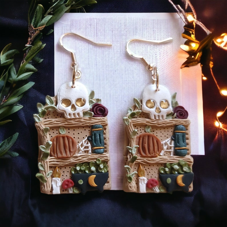 K&ouml;nnte beinhalten: Ein Paar goldene Ohrh&auml;nger mit einem Miniatur-Design aus Holzregalen. Jedes Regal ist mit einem Sch&auml;del, einem K&uuml;rbis, einem Kessel und anderen Halloween-Dekorationen verziert.